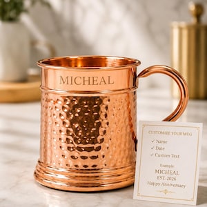 Puede incluir: Una taza de metal martillado color cobre con un asa curva. El nombre MICHEAL está grabado en el borde superior. Una tarjeta blanca está colocada al frente con el texto: CUSTOMIZE YOUR MUG, Name, Date, Custom Text, Example: MICHEAL, EST. 2026, Happy Anniversary.