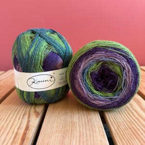 Peut inclure: Deux pelotes de fil Kauni aux tons verts, bleus et violets reposent sur une surface en bois. Le fil est enroulé en forme de boule, avec une étiquette blanche autour d'une des boules. L'étiquette porte le logo Kauni et d'autres textes.