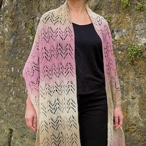 May include: A long, open-front knitted shawl in a chevron pattern. The shawl transitions from a light beige to a soft pink hue. The woman in the image is wearing a black top and pants. The background is a stone wall.