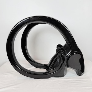 May include: A high-gloss black sculpture with a contemporary design. The artwork features a large, curved loop connected to a stylized animal form, possibly a ram or goat. The piece has a polished finish and is displayed on a white surface.