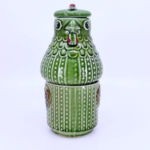 May include: A green ceramic bird-shaped container with a lid. The container is composed of two parts, a base and a top, with a bird's face design on the top. The container has a textured surface with vertical lines and white dots. The lid has a small red accent.