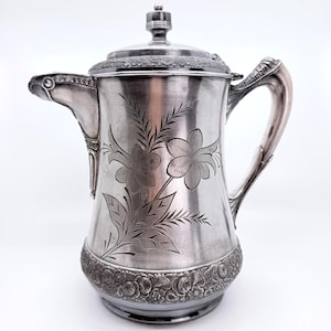 May include: A silver-toned metal coffee pot featuring an engraved floral design on the body, a textured rose pattern along the base and lid rim, a curved handle, and a spout. The lid is topped with a small, rounded finial knob.