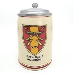 以下が含まれることがあります： 金属製の蓋が付いたクリーム色の陶器製ビアジョッキ。前面には、黄色の縦縞、赤色の側面、翼が付いた交差する稲妻が描かれた盾のエンブレムがあります。テキストには「5./12」、「5./Fm Rgt 12」、「GEISSBÜHL」と記されています。