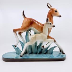 May include: A ceramic figurine of two deer, a doe and a fawn, in a running pose. The doe is brown with white legs, and the fawn is cream-colored with brown spots. They are set against blue stylized grass and a blue base.