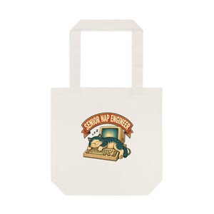 May include: A cream-colored canvas tote bag featuring a graphic of a tabby cat sleeping on a vintage computer keyboard. Above the cat, a curved orange banner displays the text SENIOR NAP ENGINEER in bold, dark-colored capital letters.