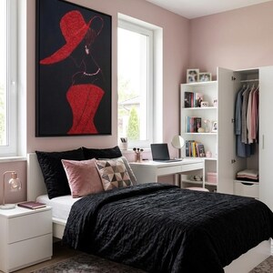 Op de afbeelding: Een slaapkamer met een zwarte doorgestikte sprei, zwarte en roze kussens, een wit bureau met een laptop, een witte boekenplank en een witte kledingkast. Een groot ingelijst kunstwerk aan de muur toont een silhouet in een rode hoed.