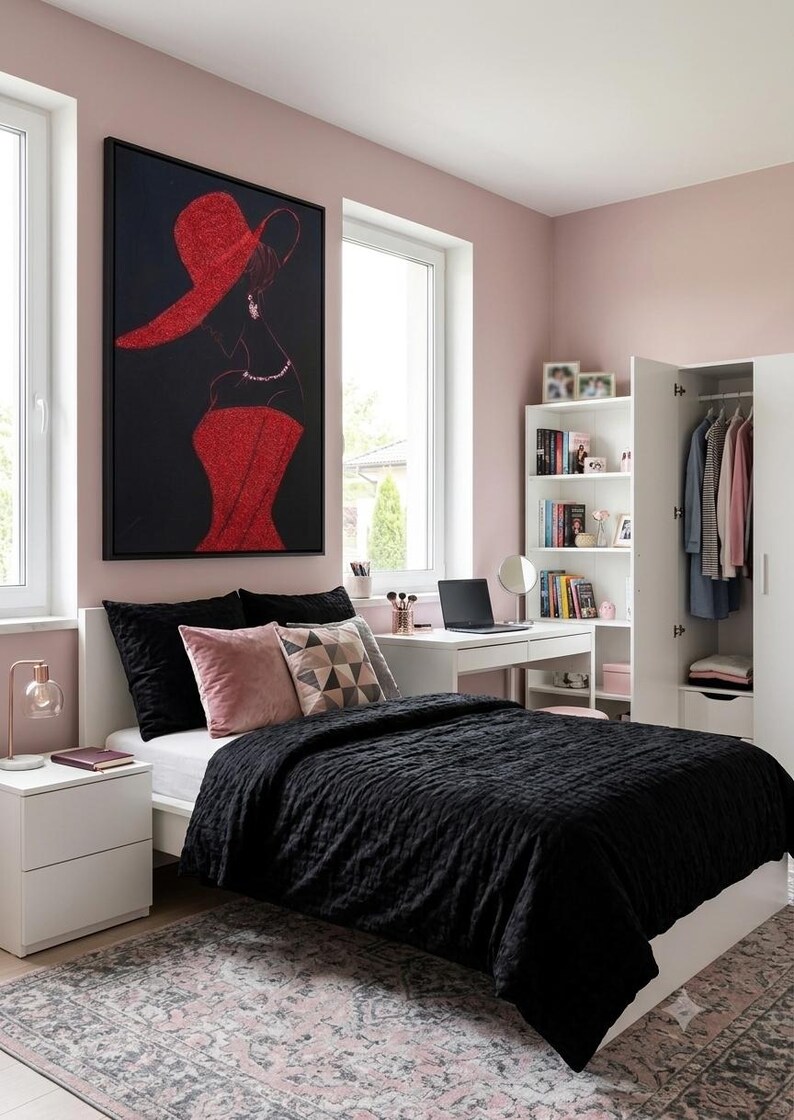 Op de afbeelding: Een slaapkamer met een zwarte doorgestikte sprei, zwarte en roze kussens, een wit bureau met een laptop, een witte boekenplank en een witte kledingkast. Een groot ingelijst kunstwerk aan de muur toont een silhouet in een rode hoed.