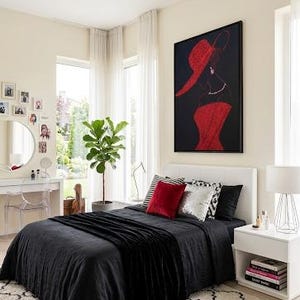 Op de afbeelding: Een rechthoekig wandkunstwerk met een gestileerde illustratie van een figuur met een rode hoed met brede rand en een rode jurk tegen een effen zwarte achtergrond. Het kunstwerk is zwart ingelijst en hangt aan een lichte slaapkamermuur.