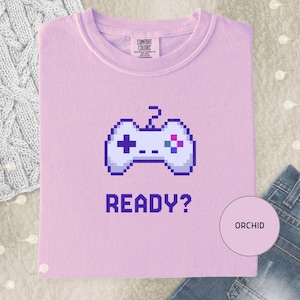 Op de afbeelding: Een lichtpaars t-shirt met een pixelachtige witte gamecontroller-afbeelding in het midden op de borst. Onder de controller staat het woord READY? gedrukt in een donkerpaars pixellettertype. Het label in de nek vermeldt Comfort Colors.