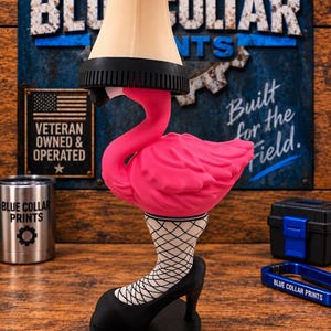 May include: A novelty lamp shaped like a bright pink flamingo with a beige lampshade resting on its head. The flamingo's neck transitions into a leg wearing a black fishnet stocking and a black high-heeled shoe, all set on a round black base.