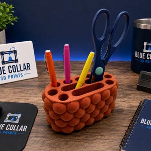 May include: An oval-shaped, burnt orange desk organizer with a textured, bubble-like exterior surface. It features multiple circular and elongated slots holding an orange pen, a pink pen, a yellow pen, and a pair of dark blue scissors.
