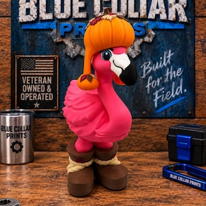 May include: A bright pink flamingo figurine wearing a pumpkin-shaped hat with autumn leaves and brown work boots. The flamingo has a black and white beak and stands upright. The figure is detailed with a textured body and realistic boot laces.
