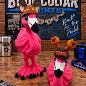 May include: Two bright pink flamingo figurines. The taller one wears a brown helmet with antlers. The shorter one wears a brown hat with two small orange pumpkin-shaped toppers. Both have white and black beaks and detailed feather textures.