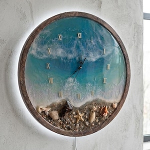Könnte beinhalten: Runde Wanduhr mit Strandmotiv. Das Zifferblatt zeigt ein blau-weißes Ozeanwellen-Design mit goldenen römischen Ziffern. Sie ist in einem braunen Holzrahmen eingefasst und enthält Muscheln und einen Seestern. Der Durchmesser beträgt ca. 30 cm.