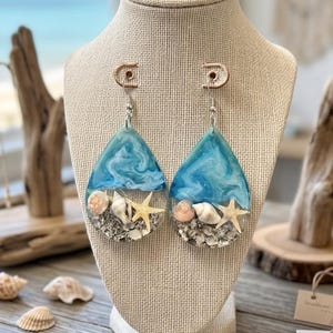 May include: Teardrop-shaped earrings with a beach theme. The top portion is a swirling blue and white design, resembling ocean waves. The bottom half features seashells, sand, and a starfish. The earrings are displayed on a beige jewelry stand.