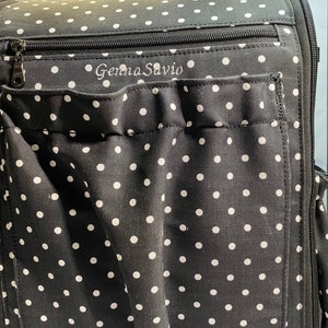 Puede incluir: Bolso tote de tela negra con lunares blancos, con cierre de cremallera en la parte superior y un bolsillo grande en la parte delantera. El bolso tiene el nombre "Gemma Savio" bordado en la parte delantera.