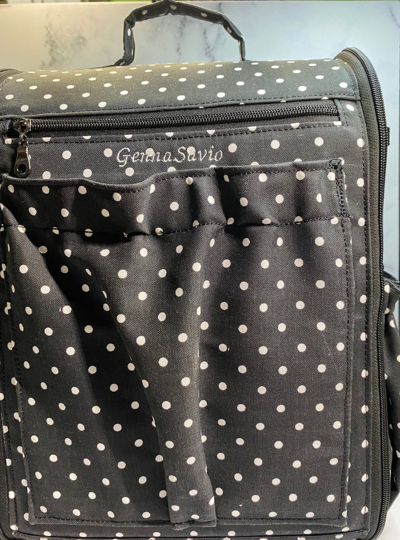 Puede incluir: Bolso tote de tela negra con lunares blancos, con cierre de cremallera en la parte superior y un bolsillo grande en la parte delantera. El bolso tiene el nombre "Gemma Savio" bordado en la parte delantera.