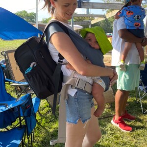 Puede incluir: Una mujer con una mochila negra y pantalones cortos de mezclilla lleva a un beb&eacute; en un portabeb&eacute;s gris. El beb&eacute; lleva un sombrero de sol verde. La mujer est&aacute; de pie en un campo de hierba.