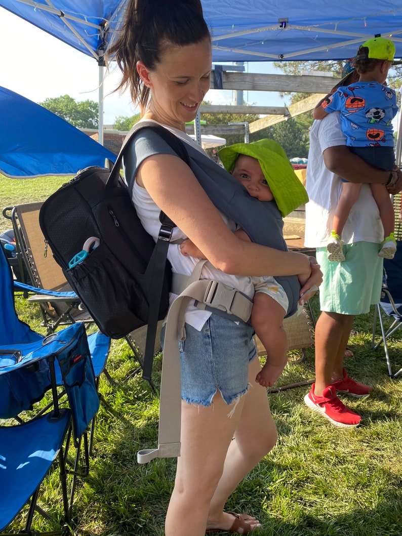 Puede incluir: Una mujer con una mochila negra y pantalones cortos de mezclilla lleva a un beb&eacute; en un portabeb&eacute;s gris. El beb&eacute; lleva un sombrero de sol verde. La mujer est&aacute; de pie en un campo de hierba.