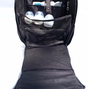 Puede incluir: Una bolsa de pa&ntilde;ales negra con m&uacute;ltiples bolsillos y un compartimento de malla. La bolsa est&aacute; abierta y hay varias botellas de beb&eacute; dentro.