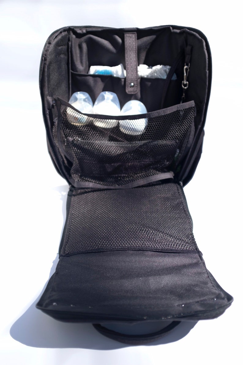 Puede incluir: Una bolsa de pa&ntilde;ales negra con m&uacute;ltiples bolsillos y un compartimento de malla. La bolsa est&aacute; abierta y hay varias botellas de beb&eacute; dentro.