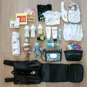 Puede incluir: Una bolsa de pa&ntilde;ales negra con una colchoneta para cambiar, un babero blanco y negro, un body blanco con estampado de animales blanco y negro, una camiseta negra con el texto "Best Cat", un libro titulado "Here Comes Fall", un libro titulado "Kelton", dos biberones transparentes, una botella de loci&oacute;n, una botella de protector solar, una cuchara para beb&eacute; azul y blanca, un mordedor azul y blanco, un contenedor de toallitas h&uacute;medas para beb&eacute; azul y blanco, un juguete para beb&eacute; azul y blanco, un juguete para beb&eacute; marr&oacute;n y blanco, una manta blanca y negra, y una pila de pa&ntilde;ales.
