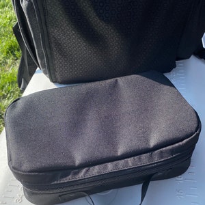 Puede incluir: Una bolsa negra con cremallera y asa. La bolsa est&aacute; hecha de un tejido resistente y tiene una superficie lisa y plana.