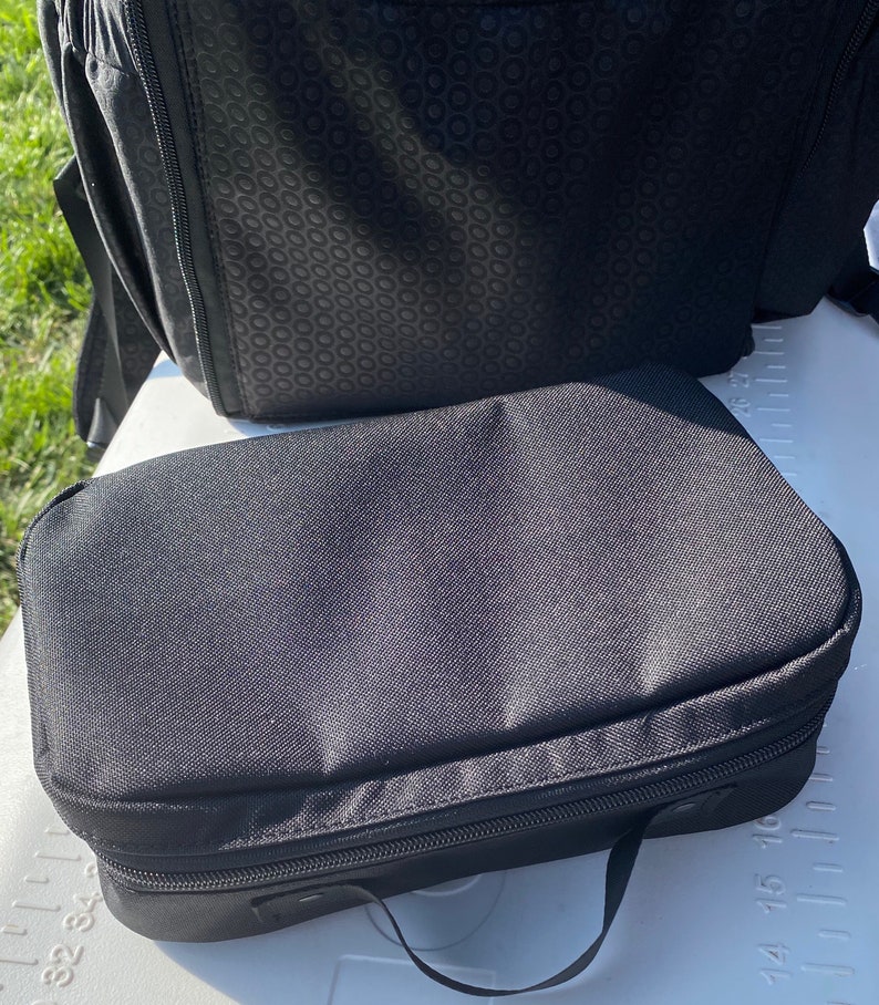 Puede incluir: Una bolsa negra con cremallera y asa. La bolsa est&aacute; hecha de un tejido resistente y tiene una superficie lisa y plana.