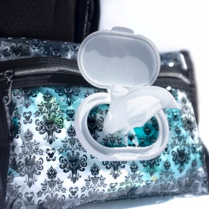 Puede incluir: Un dispensador de toallitas h&uacute;medas de viaje de pl&aacute;stico transparente con un patr&oacute;n de Haunted Mansion en blanco y negro. El dispensador tiene una tapa blanca que est&aacute; abierta, revelando una toallita h&uacute;meda blanca.