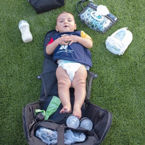 Puede incluir: Una bolsa de pa&ntilde;ales negra con un beb&eacute; acostado dentro. La bolsa est&aacute; abierta y contiene varios art&iacute;culos para beb&eacute;s, como un biber&oacute;n, toallitas h&uacute;medas y un pa&ntilde;al. El beb&eacute; lleva una camisa azul y blanca y un pa&ntilde;al.