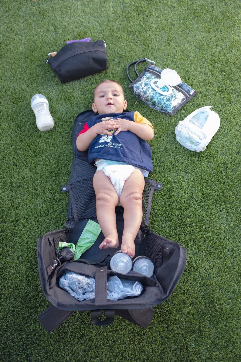 Puede incluir: Una bolsa de pa&ntilde;ales negra con un beb&eacute; acostado dentro. La bolsa est&aacute; abierta y contiene varios art&iacute;culos para beb&eacute;s, como un biber&oacute;n, toallitas h&uacute;medas y un pa&ntilde;al. El beb&eacute; lleva una camisa azul y blanca y un pa&ntilde;al.