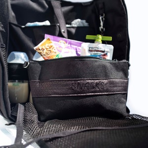 Puede incluir: Una bolsa de pa&ntilde;ales negra con una bolsa con cremallera etiquetada como "SNACKS". La bolsa contiene una bolsa de nueces, una bolsa morada y una bolsa de comida para beb&eacute;s verde y blanca etiquetada como "KIRKLAND SIGNATURE ORGANIC".