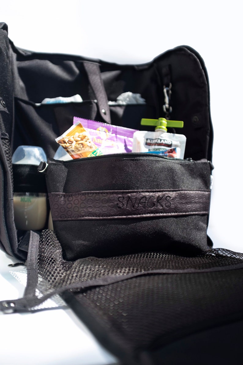 Puede incluir: Una bolsa de pa&ntilde;ales negra con una bolsa con cremallera etiquetada como "SNACKS". La bolsa contiene una bolsa de nueces, una bolsa morada y una bolsa de comida para beb&eacute;s verde y blanca etiquetada como "KIRKLAND SIGNATURE ORGANIC".