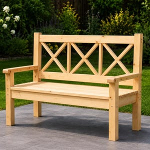 May include: A light-colored wooden bench with armrests and a decorative back featuring an X-shaped design. The bench is outdoors on a patio, surrounded by green grass and foliage.