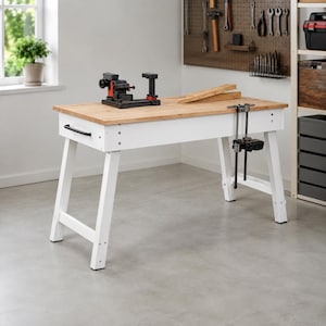 May include: A white workbench with a light wood top, featuring a black metal handle on the side, a black mechanical vise mounted on the surface, and a vertical metal clamp attached to the front edge, set against a wall with a pegboard tool organizer.
