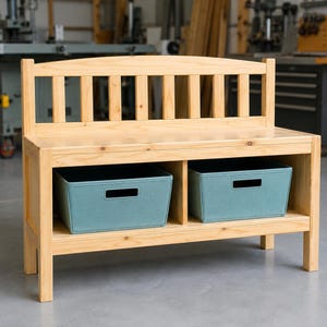 Puede incluir: Un banco de madera de color claro con respaldo de listones y asiento plano. La base incluye dos compartimentos de almacenamiento abiertos, cada uno con una caja de tela rectangular de color verde azulado con un asa recortada al frente.
