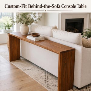 May include: A long, rectangular console table made of medium-toned wood with a smooth surface and two vertical side supports. The table is positioned directly behind a cream-colored sofa. The text Custom-Fit Behind-the-Sofa Console Table is visible.