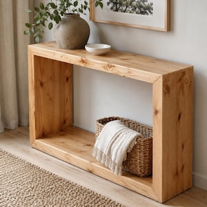 Op de afbeelding: Een rechthoekige lichtbruine houten sidetable met zichtbare nerven en knoesten. Bovenop staat een grote grijze keramische vaas met groen blad en een kleine witte kom. Onderin staat een gevlochten mand met een witte deken met franjes.