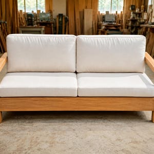 May include: A two-seater wooden sofa with a light brown frame and two white cushions. The sofa has a simple, modern design and is suitable for indoor use. The sofa is made of wood and fabric.