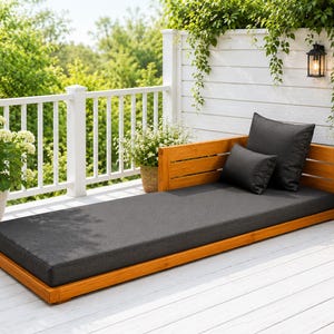 May include: A low-profile daybed constructed from warm-toned natural wood featuring a slatted backrest and side panel. The frame supports a long, rectangular charcoal gray cushion accompanied by one square and one rectangular matching pillow.