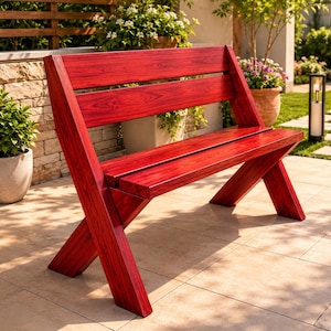 May include: A red wooden bench with an X-shaped leg design and a slatted back. The bench is outdoors on a tiled patio, surrounded by potted plants and greenery. The bench is stained a deep red color.