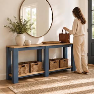 May include: A long rectangular console table with a blue frame and a light wood top. The table features a lower shelf holding four woven wicker storage baskets. A large round mirror hangs above the table, which holds a vase with greenery.