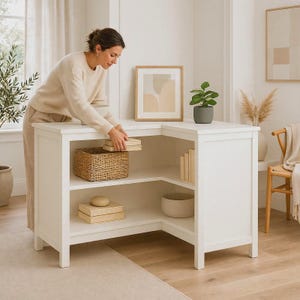 May include: A white L-shaped corner desk featuring two open shelves. The top shelf holds a woven basket and a stack of books, while the bottom shelf contains a smooth round object, a stack of books, and a large bowl. The desk has a clean finish.