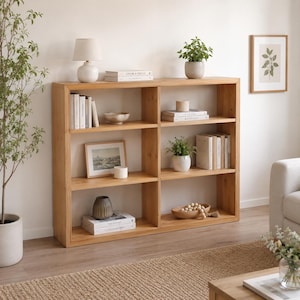 May include: A light wood bookshelf with six rectangular compartments. The shelves are decorated with white books, a small white lamp, potted green plants, a framed picture, a ceramic bowl, a dark vase, and a wooden bowl containing wooden beads.