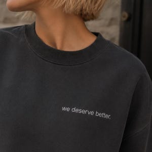 May include: Dark grey sweatshirt featuring the white text "we deserve better". The crew neck top has a simple, solid design. The slogan is embroidered across the chest in a minimalist style.