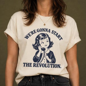May include: A cream-coloured short-sleeve t-shirt featuring a dark blue graphic of a young child holding a vintage telephone receiver. The text above and below the illustration reads, "WE'RE GONNA START THE REVOLUTION." The shirt has a crew neckline.
