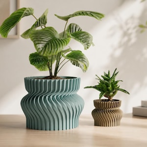 Op de afbeelding: Twee decoratieve bloempotten met planten. De grotere pot is blauwgroen met een golvend, verticaal ontwerp en de kleinere is bruin. De planten hebben groene bladeren. De potten staan op een houten oppervlak.