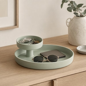 Può includere: Un vassoio verde salvia a due livelli con una base rotonda e una parte superiore più piccola ed elevata. Il vassoio contiene un portafoglio, occhiali da sole, chiavi e un orologio. Un vaso decorativo con verde è sullo sfondo.