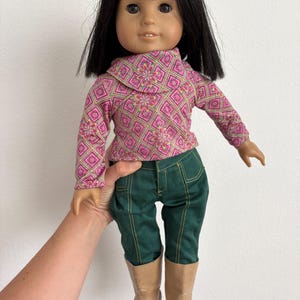 May include: A doll with black hair, wearing a pink and green patterned top, green pants, and tan boots. The doll is being held by a person.