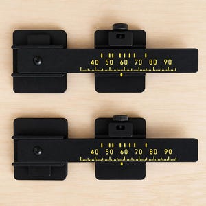 Può includere: Due strumenti di misurazione in metallo nero con segni gialli e numeri da 40 a 90. Gli strumenti hanno una base rettangolare e un meccanismo scorrevole. Gli strumenti sono posizionati su una superficie di legno chiaro.